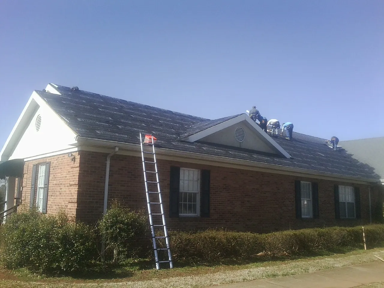 Crew tearing shingles off a brick ranch roof with a ladder set — Metal Roof Repair work by our crew in Prides Crossing