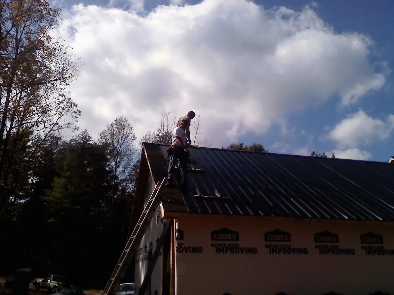 Roofer fastening dark metal panels high against a cloudy sky, from a Roof Inspection job in Prides Crossing